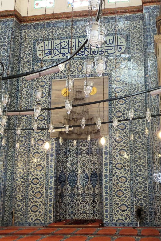 A tall carved stone mihrab stands within a wall densely covered in blue-and-white floral tiles and inscriptions, crossed by lamps and cables.