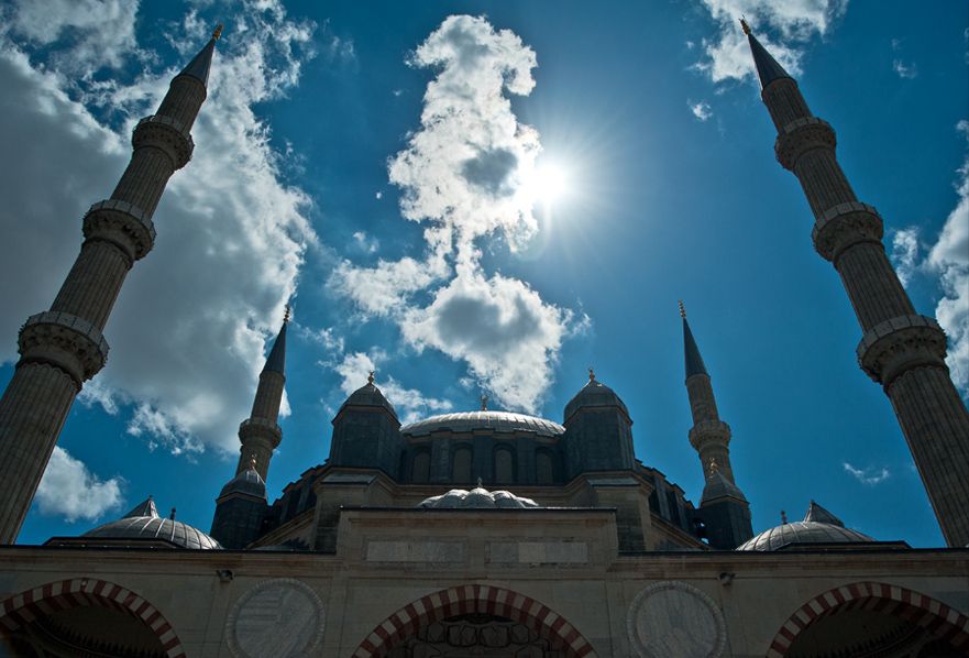 Four very tall slender minarets frame a large central dome above a shaded stone courtyard under sunlit clouds.