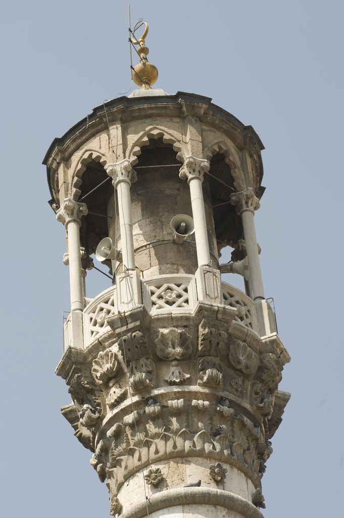 Several horn loudspeakers and cables are fixed around an ornate open stone minaret balcony beneath its finial.