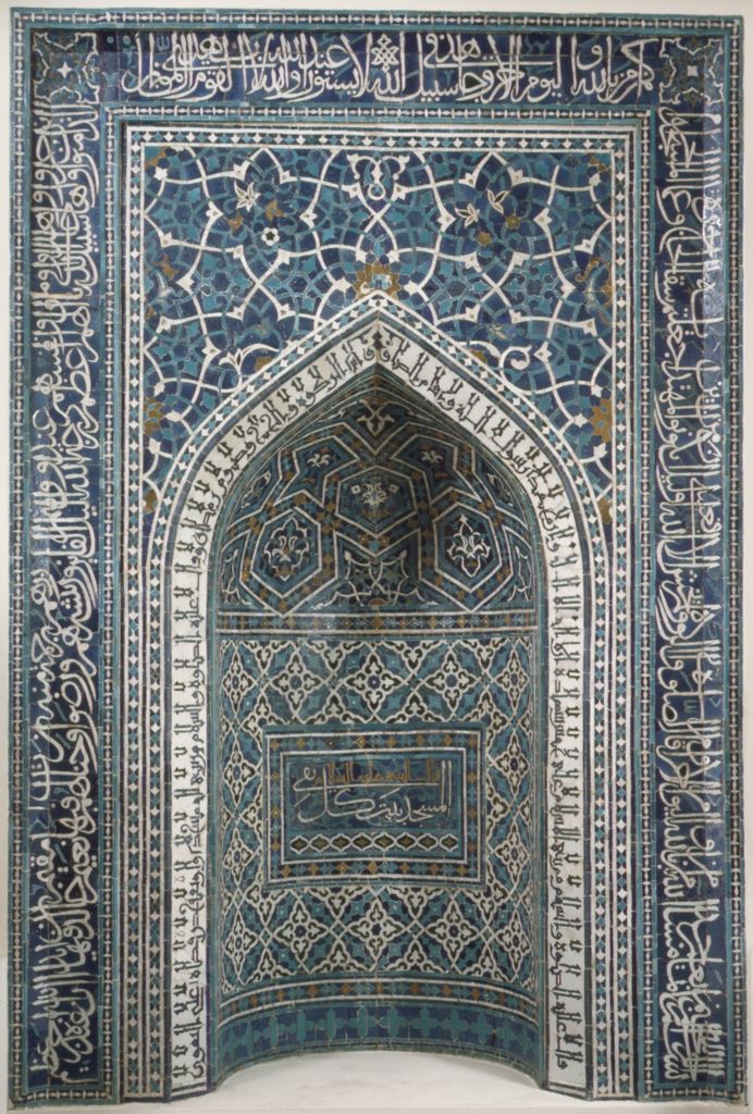 A tall pointed niche covered in blue, turquoise and white mosaic tile and calligraphy stands isolated against a pale museum background.