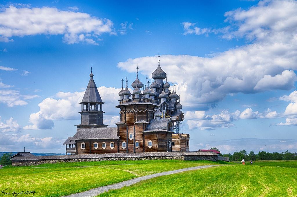 Two elaborate timber churches with many shingled domes and a wooden bell tower rise behind a low enclosure in a green lakeside landscape.