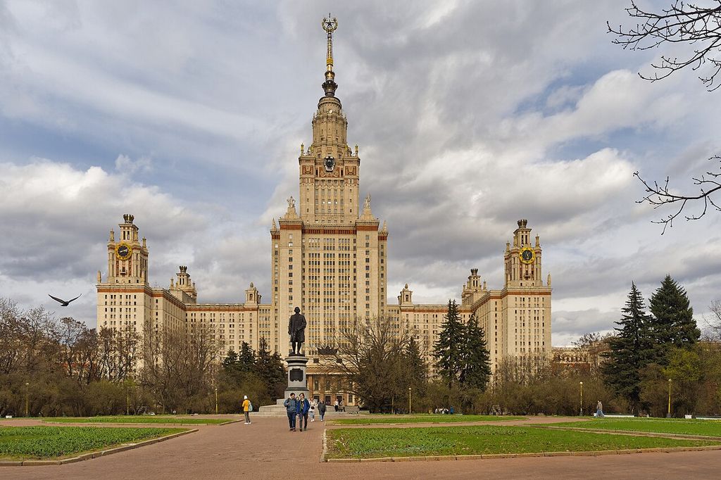 A tall symmetrical university tower with a gilded spire rises above stepped wings, corner towers, clocks, gardens and a central approach.