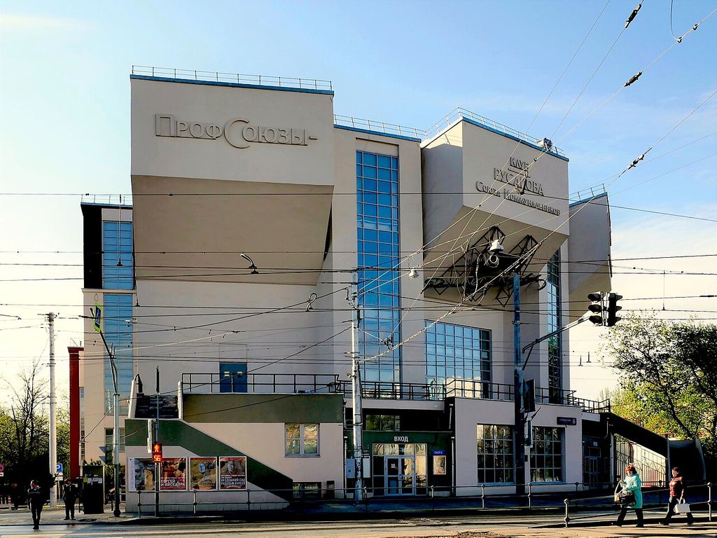 A pale angular club building has three large projecting upper volumes, tall blue-glazed strips and steep exterior stairs above a busy street corner.