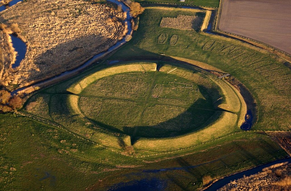 An aerial view shows a circular grass-covered earthwork with four entrances, crossing paths and rectangular building traces among fields and waterways.