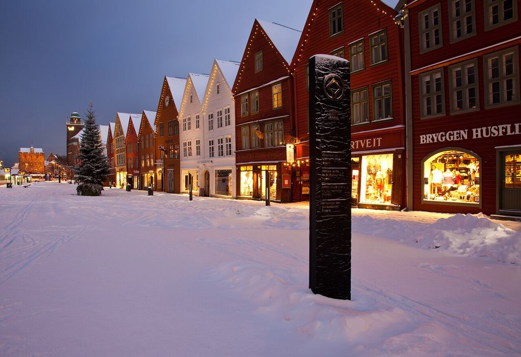 A row of narrow red, white and ochre timber buildings with steep gables and lit shop windows lines a broad snow-covered street at dusk.