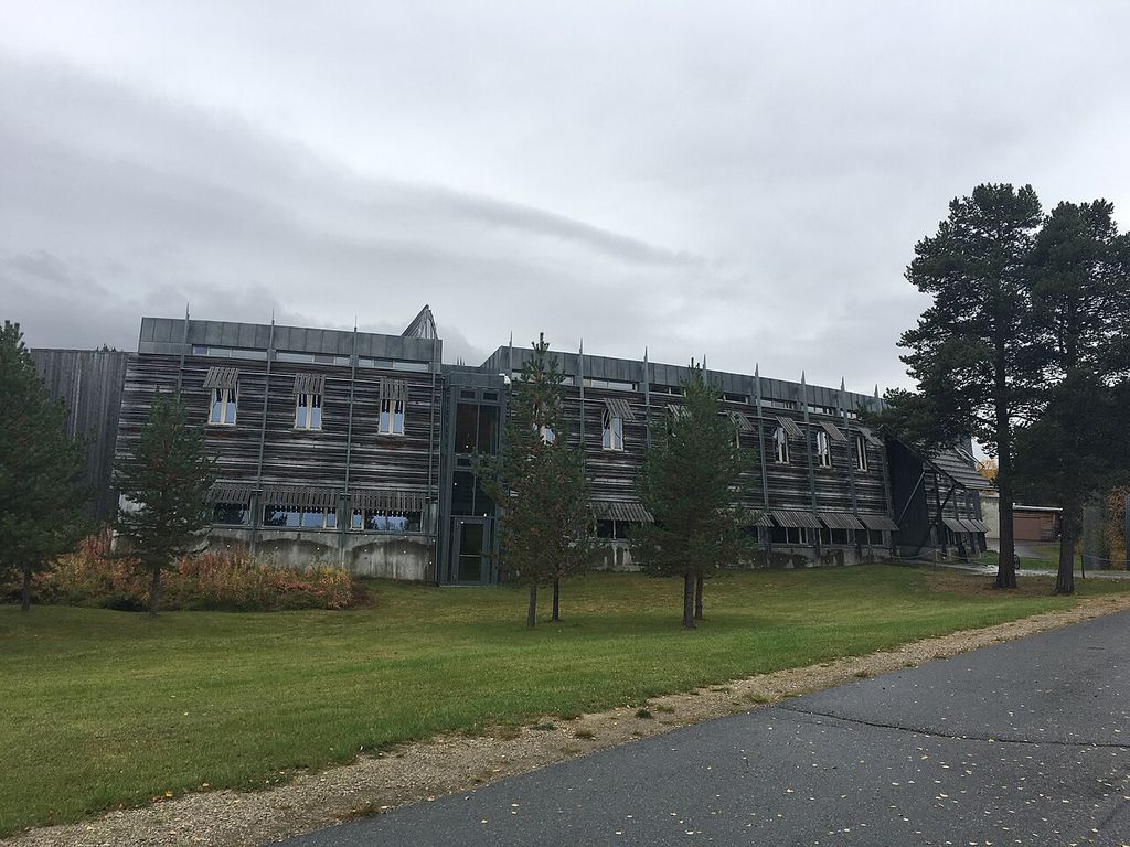 A long low civic building with horizontal timber screens, glass entrances and a taller curved roof volume stands among grass and pine trees under a grey sky.