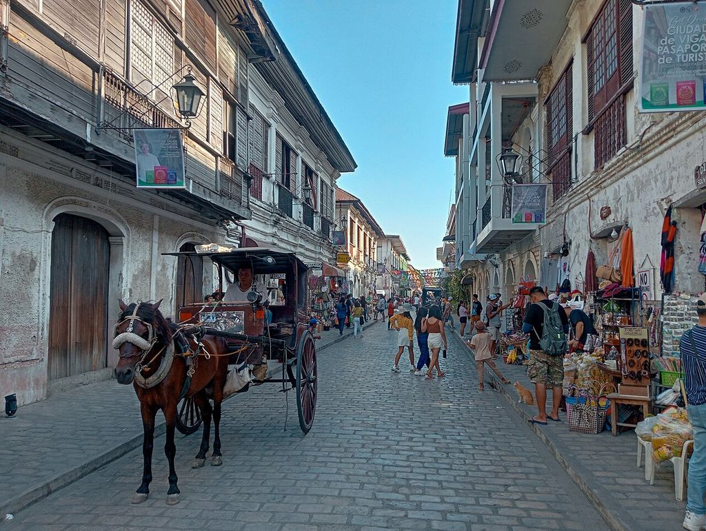 Closely spaced buildings with timber upper storeys and street-level shops line a paved street with visitors and a horse-drawn carriage.