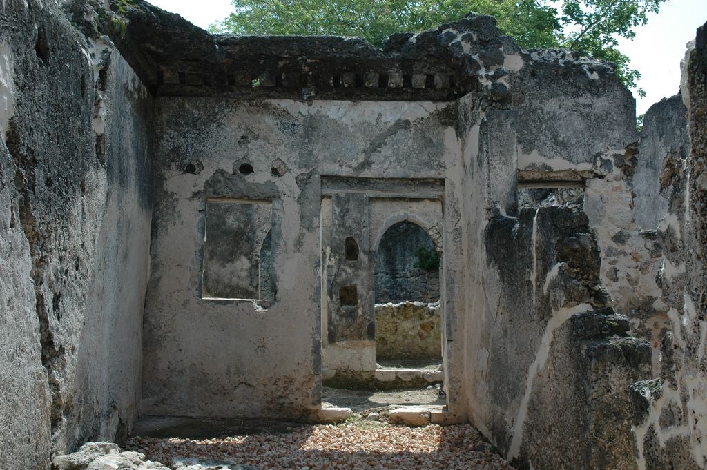 A roofless sequence of small plastered stone rooms has aligned rectangular doorways and windows, with rubble underfoot and vegetation above the walls.
