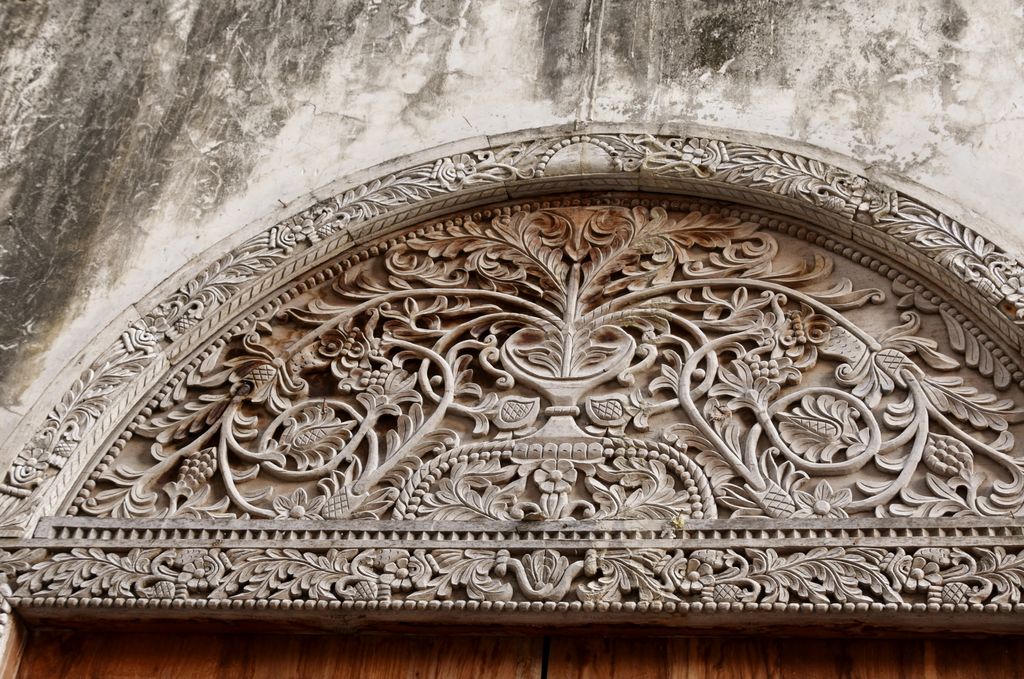 A semicircular wooden transom and surrounding bands are densely carved with curling leaves, flowers and a central vase-like motif.
