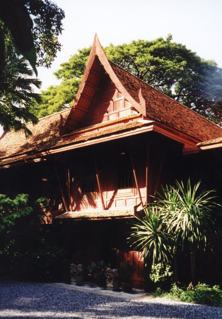 A dark red raised timber house with a steep tiled roof and deep eaves stands among dense tropical plants.