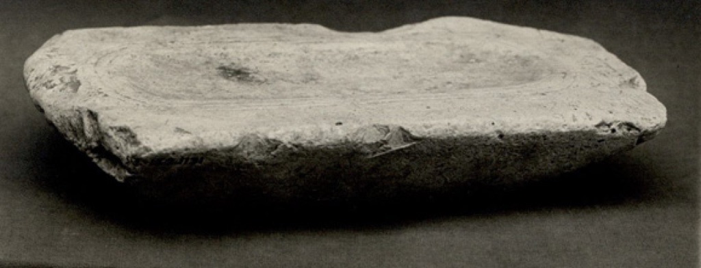 A small worn rectangular limestone painting palette lies against a dark background, its shallow working surface visible from a low angle.