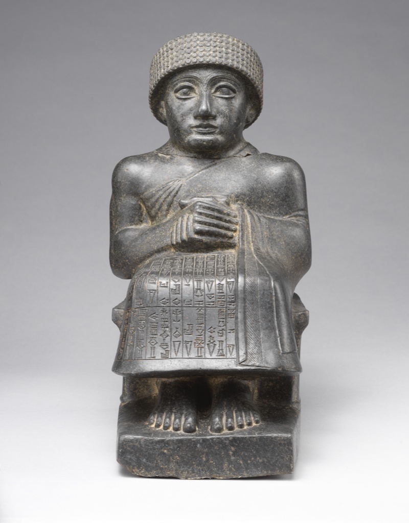 A dark diorite statue of seated ruler Gudea has folded hands, a patterned cap and a cuneiform-covered robe.